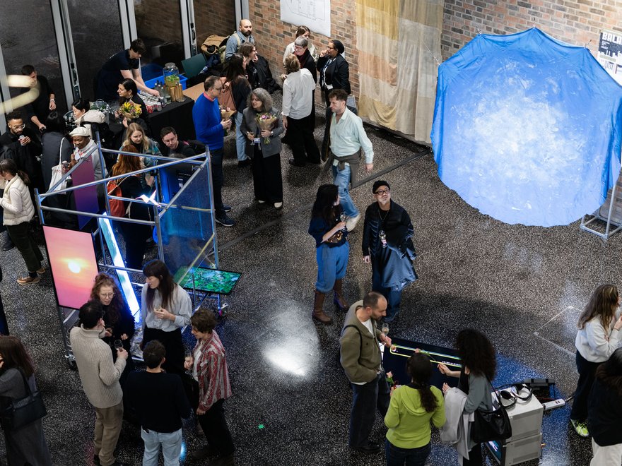 Overhead view of a crowded indoor art exhibition or event with people mingling and interacting with various installations, including a large glowing blue structure and illuminated screens.