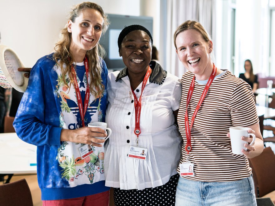 Three smiling women stand together, two holding mugs, all wearing lanyards with "Royal College of Art" visible on them.
