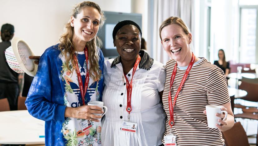 Three smiling women stand together, two holding mugs, all wearing lanyards with "Royal College of Art" visible on them.