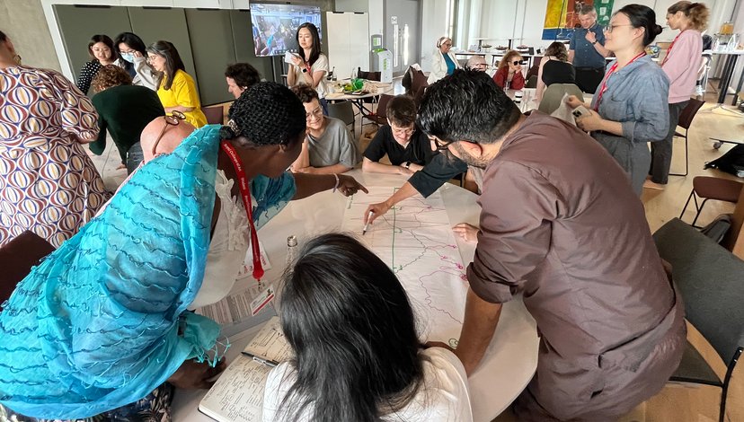 A high-angle view of an interactive workshop session where participants are using markers to annotate a large paper master plan. A woman in a vibrant blue scarf points to a specific area of the map while others look on intently.