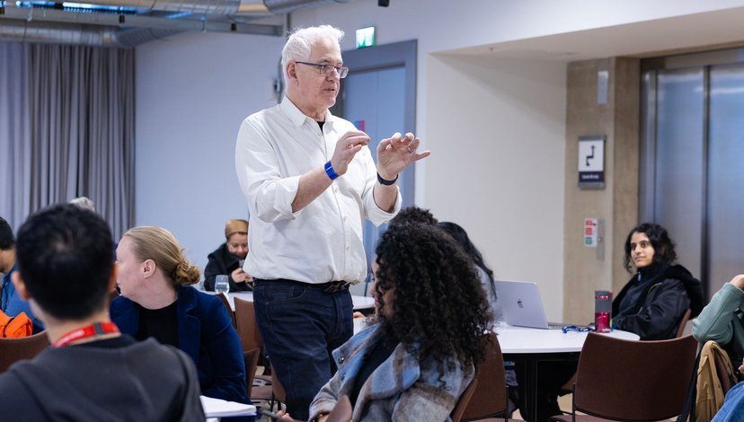 A white-haired man in a white shirt and dark jeans stands and gestures with both hands while speaking to a group of workshop participants seated at round tables.