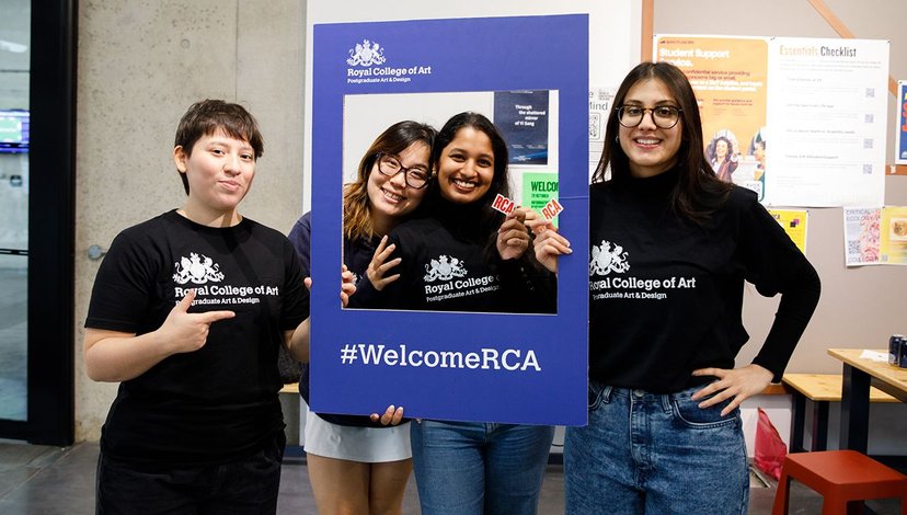Four students wearing Royal College of Art t-shirts pose with a #WelcomeRCA frame at a welcome event, smiling and cheerful.