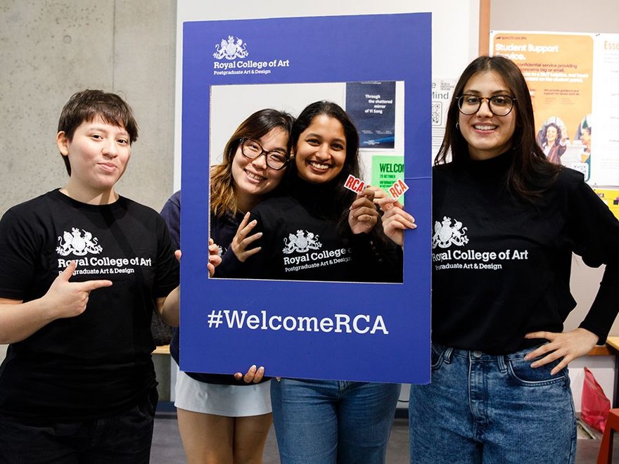 Four students wearing Royal College of Art t-shirts pose with a #WelcomeRCA frame at a welcome event, smiling and cheerful.