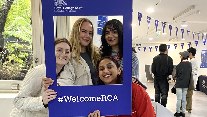 Four smiling students posing together inside a large blue photo frame with the text ‘Royal College of Art – Postgraduate Art & Design’ at the top and the hashtag ‘#WelcomeRCA’ at the bottom